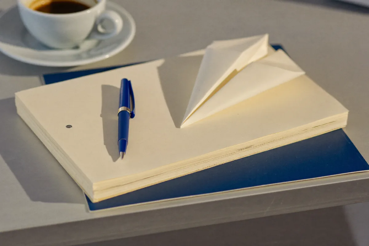 Stapel leerer Papierbögen mit gefaltetem Papierflieger, marineblauem Stift und Espresso auf einem Schreibtisch im Morgenlicht